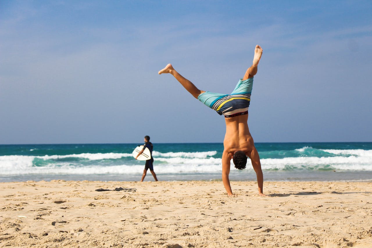 services-01 Man doing a handstand on a sunny beach with surfer in background, evoking a sense of freedom and joy.